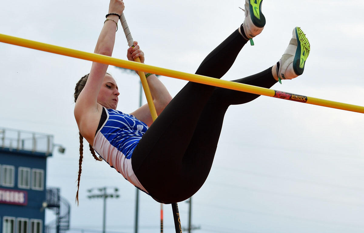 Female athlete in a blue and white uniform vaulting over a yellow bar during a pole vault event outdoors.