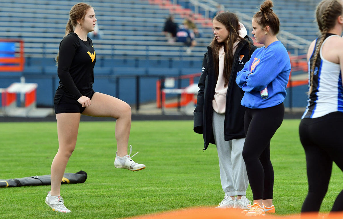 Young woman in black athletic wear lifts her knee as others watch on a track field.
