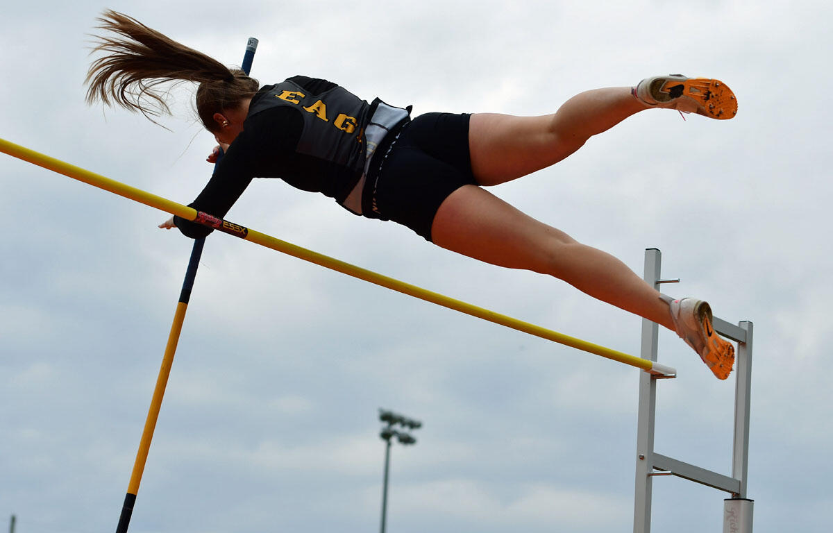 Female pole vaulter in mid-air clearing a yellow bar with a pole, wearing a black uniform and orange shoes.