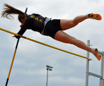 Female pole vaulter in mid-air clearing a yellow bar with a pole, wearing a black uniform and orange shoes.