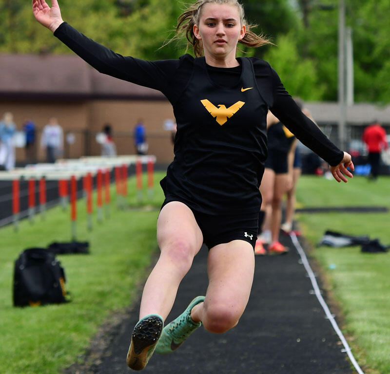 Female athlete in black uniform with yellow logo mid-air during a long jump at an outdoor track meet, with green field in the background.