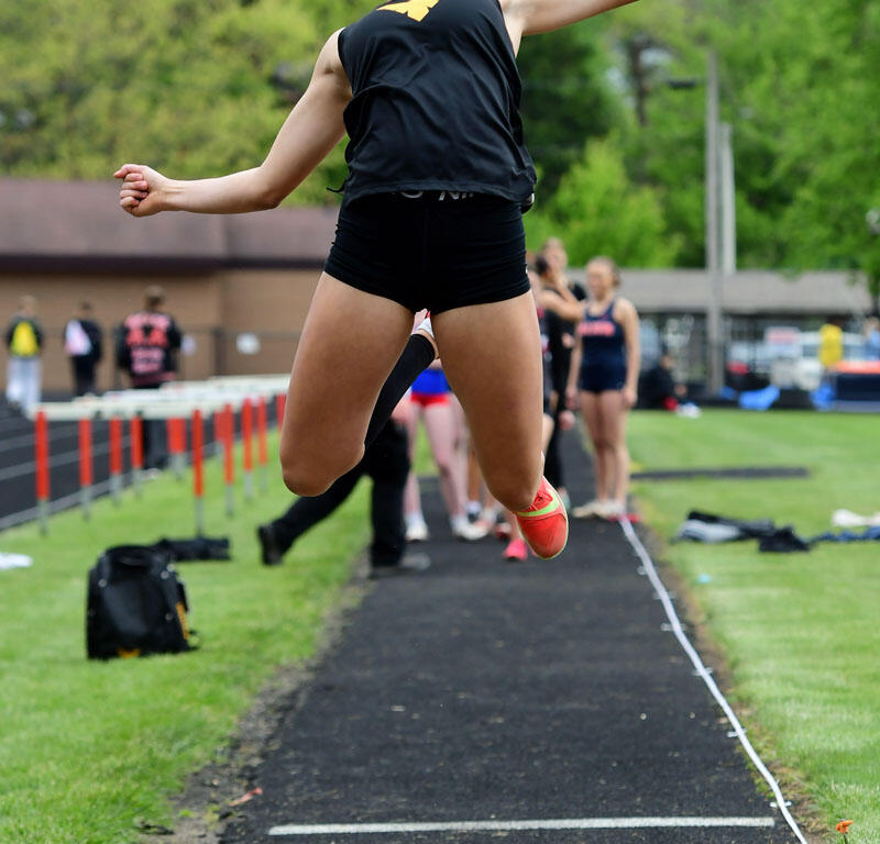 Female athlete in a black tank top jumps mid-air during a long jump on a track, sand pit visible ahead and bystanders in the background.