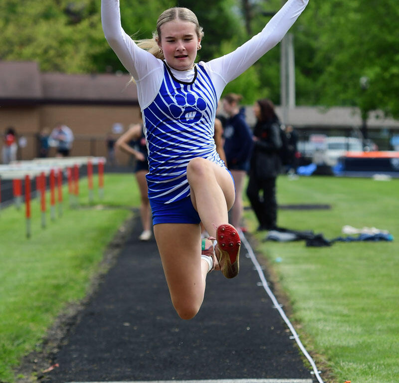 Young female athlete in a blue and white uniform leaps mid-air during a long jump on a track, arms raised and legs extended.
