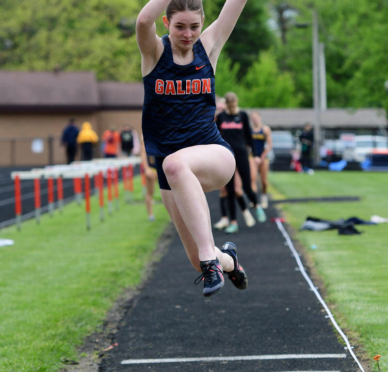 Female track and field athlete in a Galion uniform mid-jump during a long jump event, leaving the sand pit behind the runway.