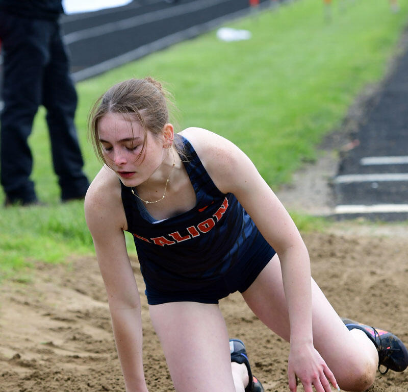 Female athlete kneeling in a sand pit during a long jump attempt at a track meet, wearing a navy uniform with orange lettering.