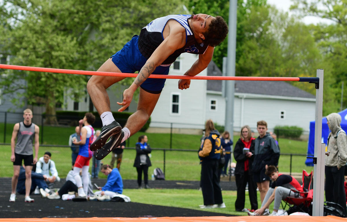 Athlete clears a high jump bar mid-air during a track meet, stadium and spectators in the background.