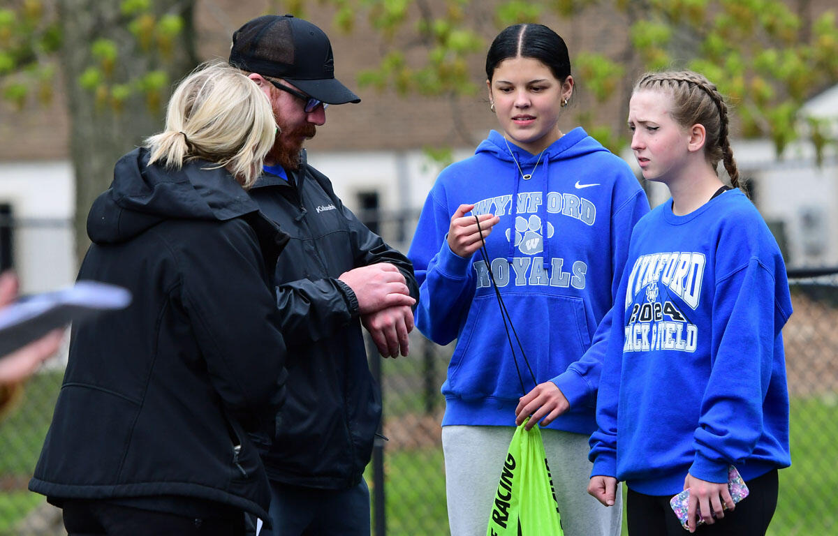 Two girls in blue hoodies labeled 'Waynf ord Royals' talk with a man in a cap outdoors, one holding a bright green drawstring bag.
