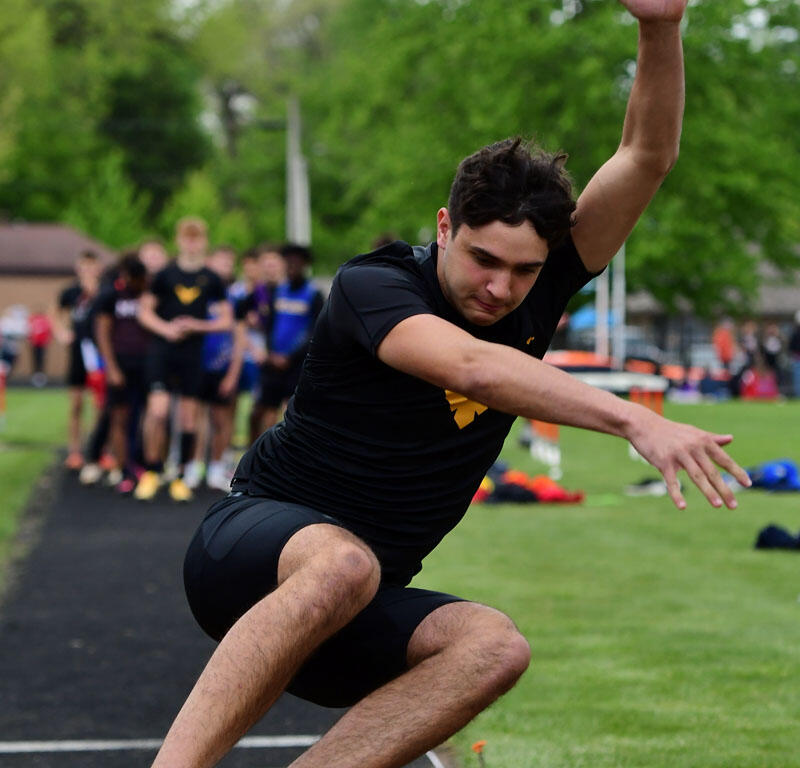 Male athlete in a black uniform mid-air during a long jump, sand pit in foreground and spectators in the background.