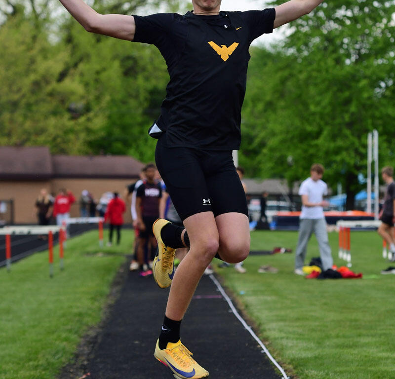 Male athlete in black shirt and yellow shoes leaps mid-air during a long jump on the track, sand pit ahead, arms outstretched.