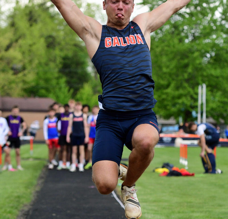 Male athlete in a Galion uniform mid-jump in a long jump event, arms spread, tongue out, with spectators in the background on a track field.