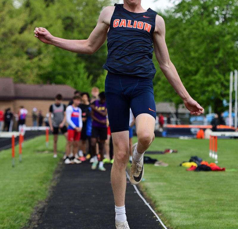 Male athlete from Galion in navy uniform mid-air during a long jump on a track, arms extended, with spectators in the background.