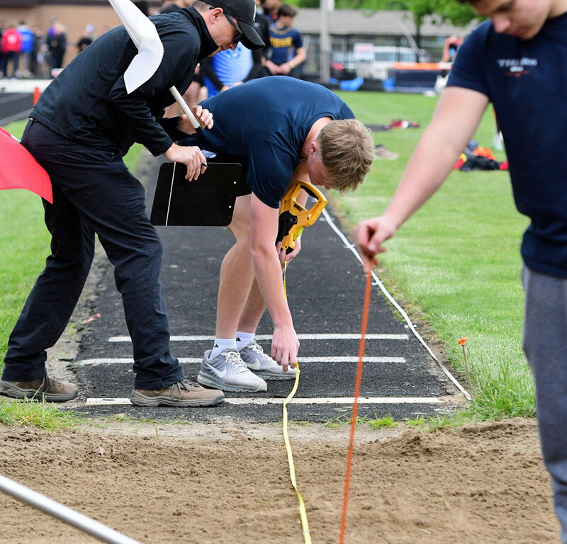 Officials measure a long jump result on a track, one holding a clipboard and another with a yellow measuring tape by the sand pit.