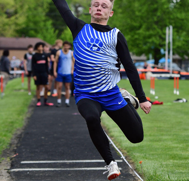 Male athlete in a blue and white striped uniform leaps mid-air in a long jump on a track, with onlookers and a grassy field in the background.