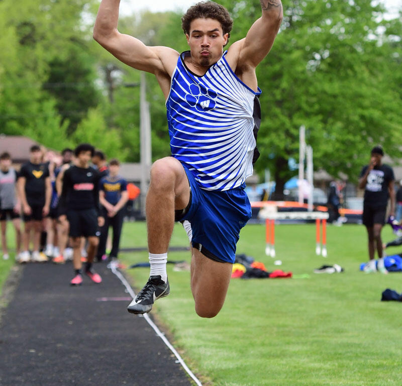 Male track athlete in a blue and white uniform mid-air during a long jump attempt on a track field.