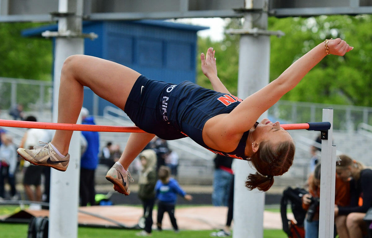 Female athlete in navy uniform on a horizontal bar, performing a backbend swing outdoors on a track field with spectators in the background.