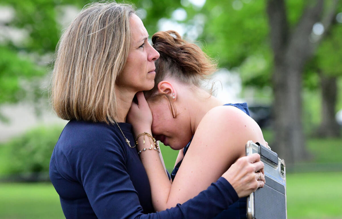 Two women in navy tops embrace in a park; one rests her head on the other's shoulder for comfort while holding a silver briefcase.