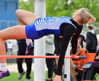 Female athlete in a blue and white striped leotard performs on a high bar during a track and field event, with spectators in the background.