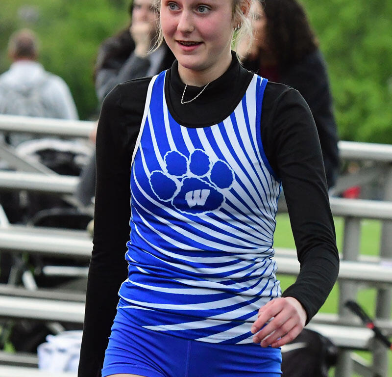 Young female athlete in a blue and white striped uniform with a paw print logo, standing outdoors on a track/field.