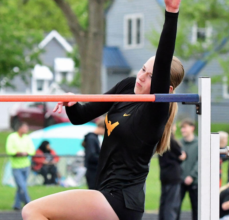 Female gymnast on an outdoor horizontal bar, reaching up with one arm while the other grips the bar.