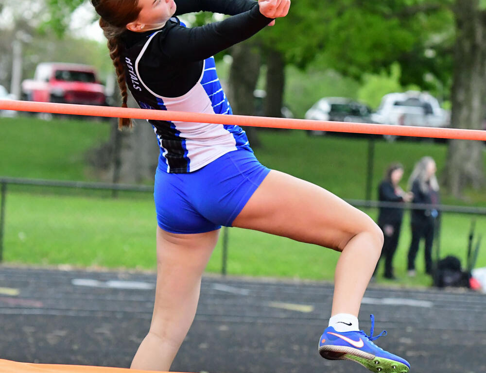 Female athlete in blue and white outfit clears an orange pole vault bar on an outdoor track field.
