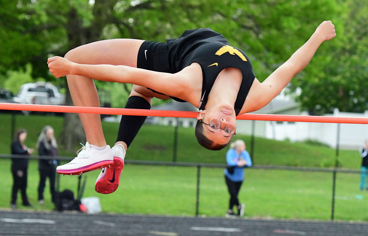 Athlete performing a backbend over a horizontal bar during an outdoor track event, wearing a black leotard and white/red spikes.