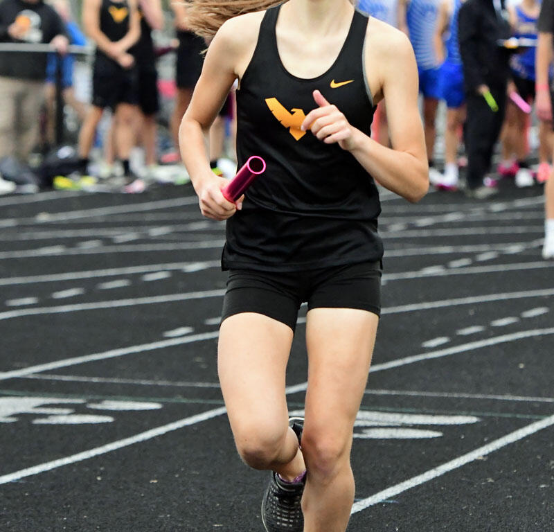 Female athlete in a black tank top sprinting on a track during a relay race, holding a pink baton with teammates behind her.