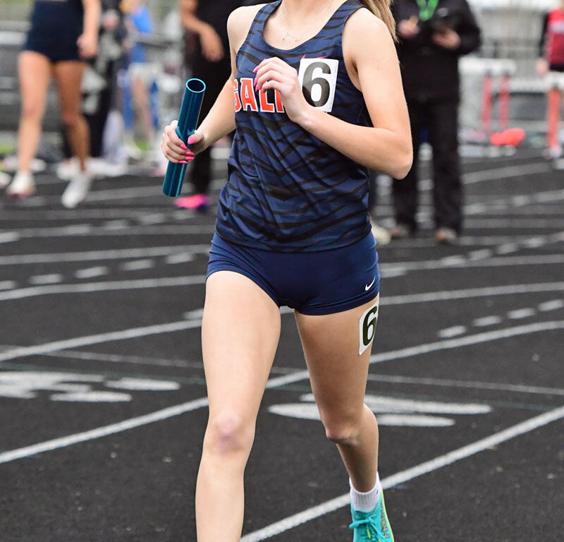 Relay runner: young woman in navy-blue uniform holds a baton while running on a track, bib number 6 visible.