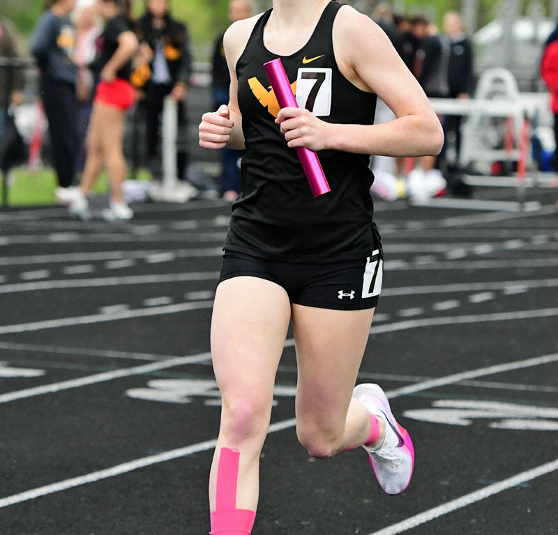 Female athlete running on a track holding a pink baton during a relay race, wearing black sportswear and pink knee tape.