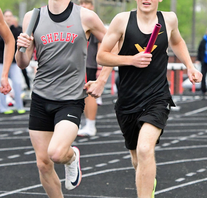 Two young male sprinters race side by side on a track, each holding a baton during a relay event.