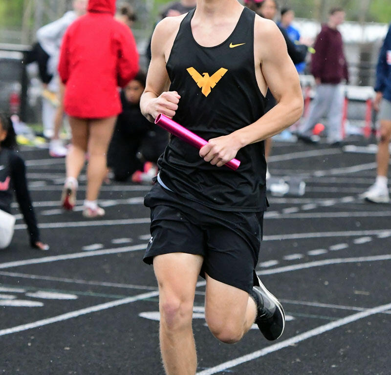 Male sprinter in a black singlet holding a pink baton during a relay on an outdoor track, with spectators in the background.