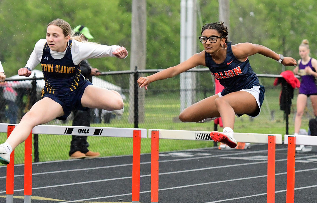 Two female hurdlers mid-race clearing hurdles on a track, one in a navy and gold uniform and the other in a navy and white uniform, rain visible in the air.