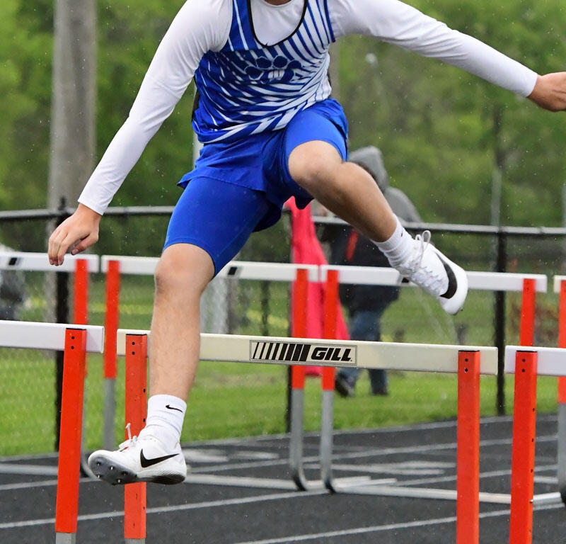 Male hurdler in a blue-and-white uniform clearing a hurdle on an outdoor track surface.