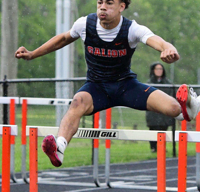 Athlete in a blue uniform jumps over orange hurdles on a track, mid-air with intense focus.