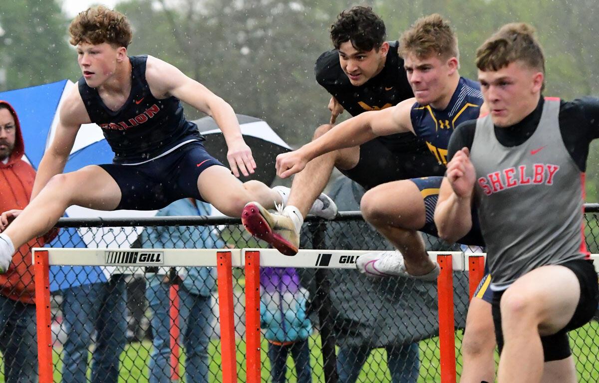 Four male runners leap over a hurdle in a track and field hurdle race, with spectators and tents in the rainy background.