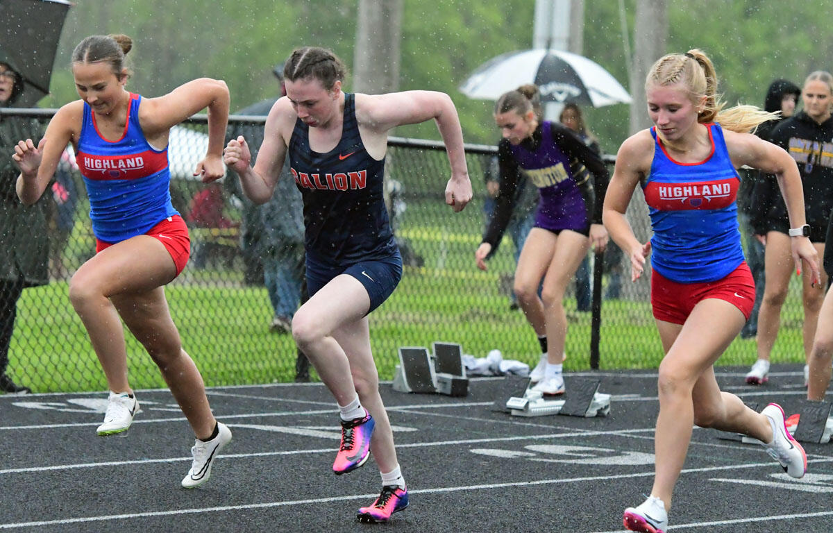 Three female sprinters push off at the start of a rainy track race; two wear Highland blue tops with red shorts, one in a navy Ballion uniform.