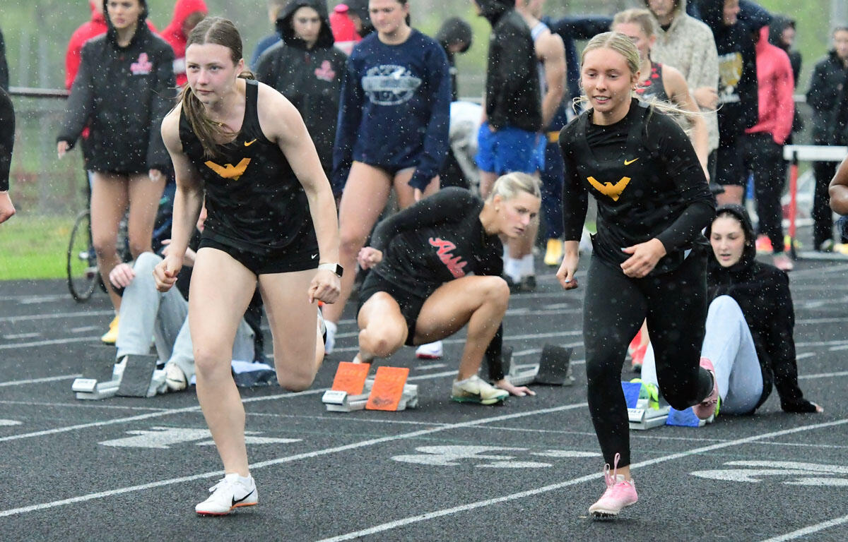 Female sprinters launch from starting blocks on a rainy track, crowds watching in the background.