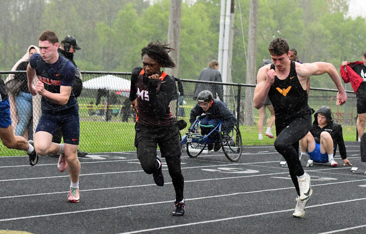 Athletes sprint on a wet track in a race, with a person in a wheelchair visible in the background under light rain behind the fence.