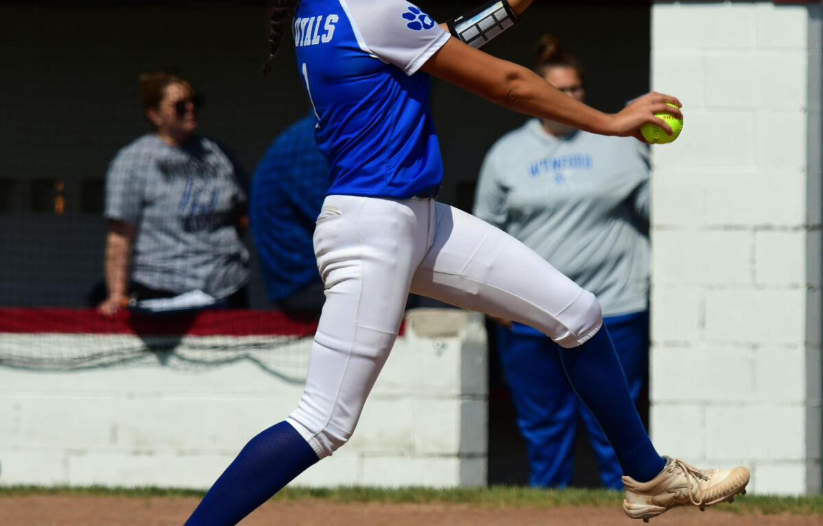 Female softball pitcher in blue and white uniform mid-throw with glove raised on a dirt field.