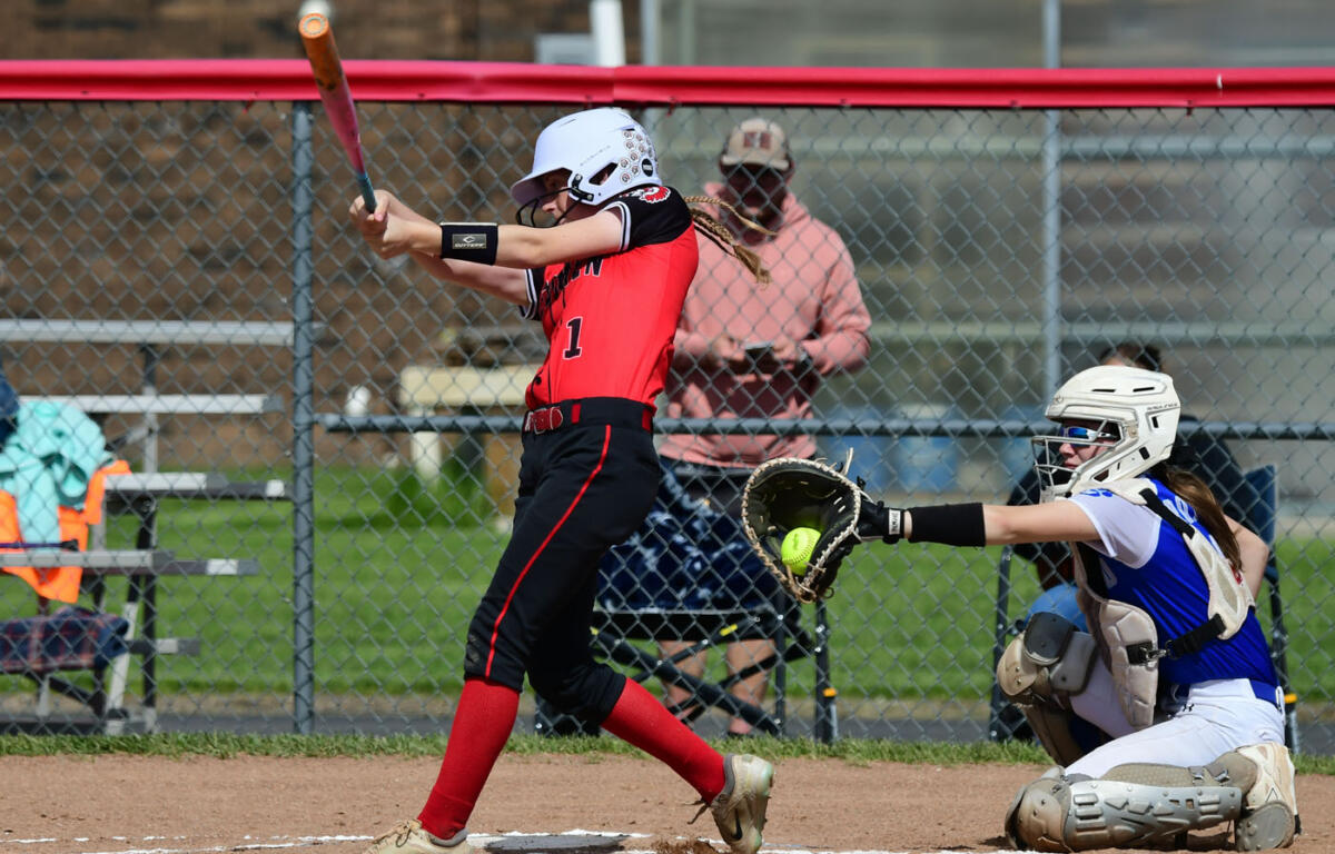 Softball batter in red and black swings at a pitch as the catcher in blue and white gear reaches with her glove near home plate behind him