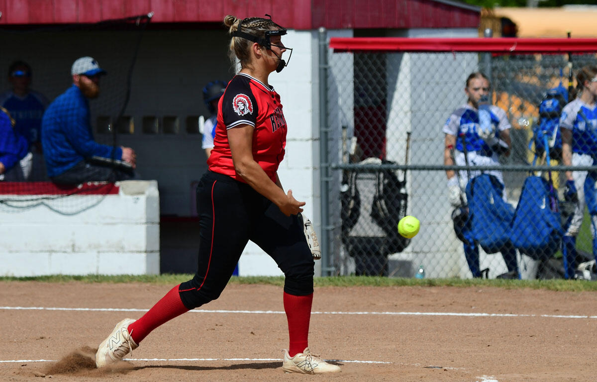 Softball pitcher in red uniform mid-pitch on a dusty infield with teammates in blue watching behind a chain-link fence in the background.