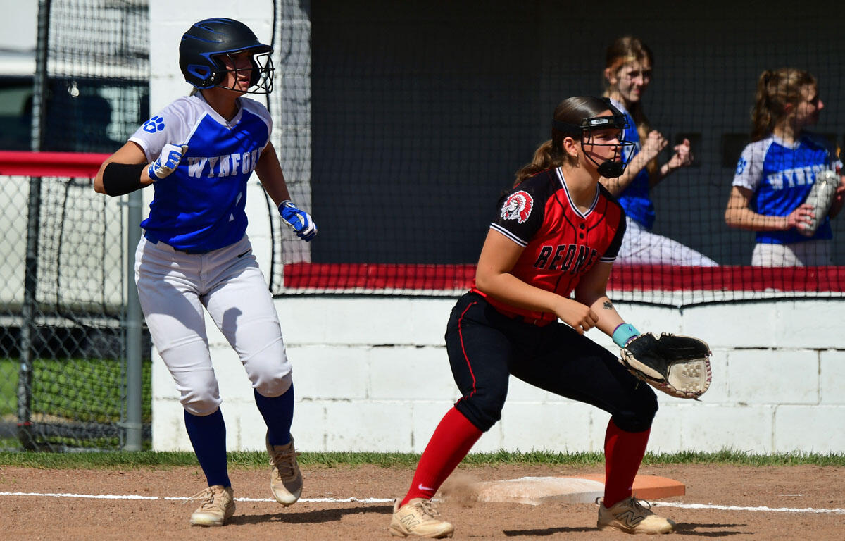 Two female softball players in blue and red uniforms near a base; blue runner heads toward the bag as the red fielder prepares to field the ball with her glove nearby behind a chain-link fence.