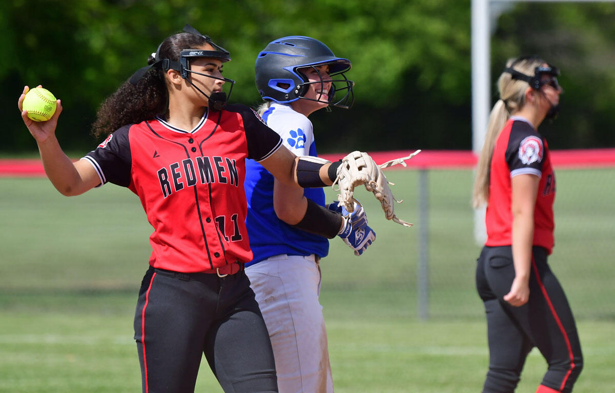 Softball players on a sunny field; a Redmen player (number 11) raises a yellow ball while catching with a glove.