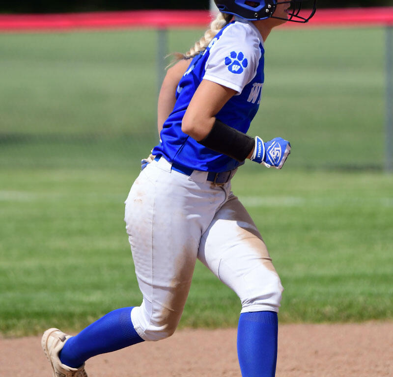 Female softball player in blue and white uniform running toward first base on a dirt infield, wearing a helmet and batting glove.
