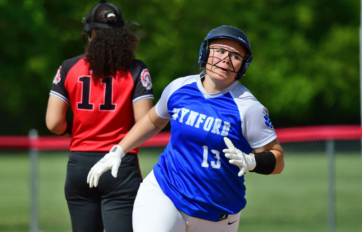 Female softball player in a blue and white Lynford jersey (number 13) sprints toward the camera while wearing a helmet and gloves on a sunny field; another player in a red jersey (number 11) faces away in the background.