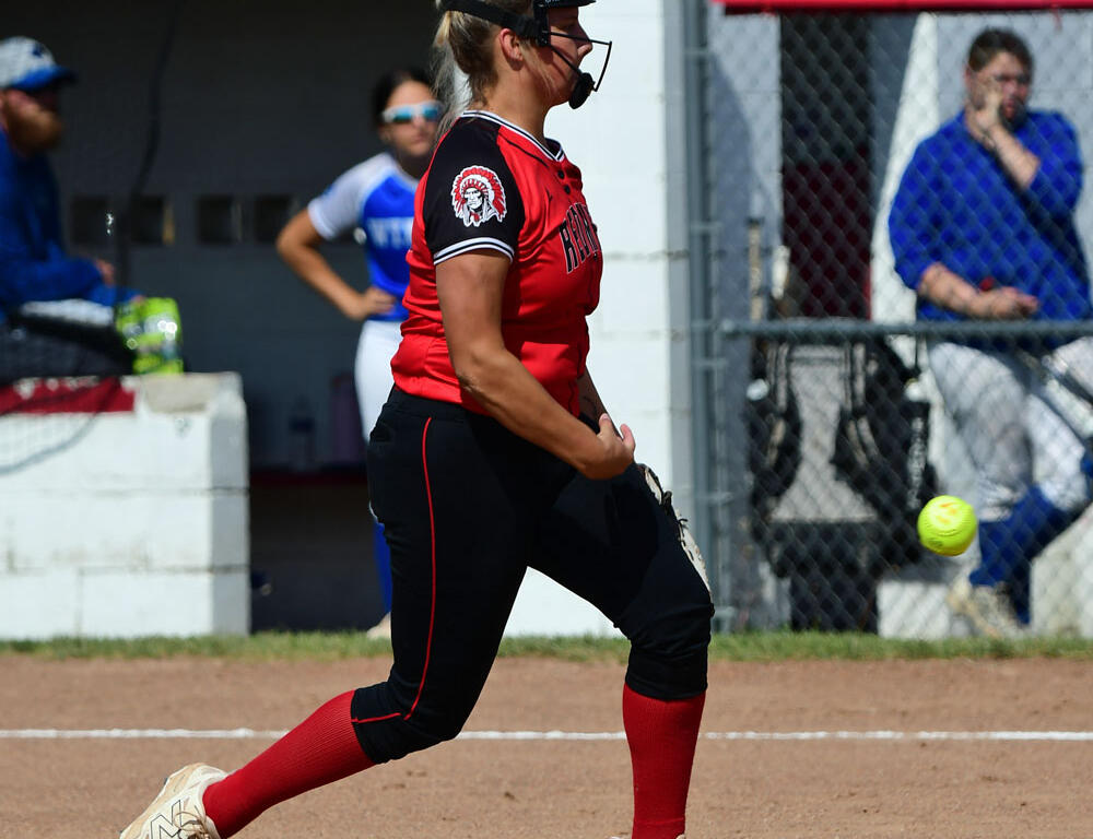 Female softball pitcher in red jersey and black pants throws a pitch on a dirt field, wearing a protective face mask and red socks.