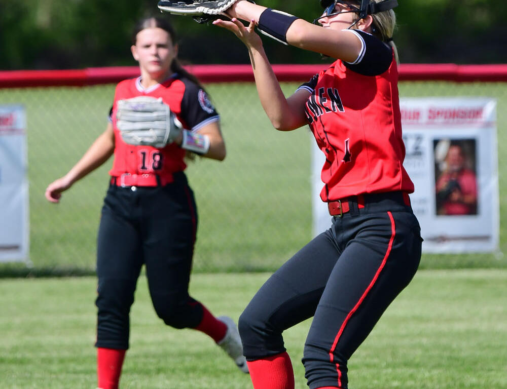 Female softball players in red and black uniforms on a grassy field; one fielder reaches with glove to catch a bright ball in midair, another teammate stands nearby in the background.