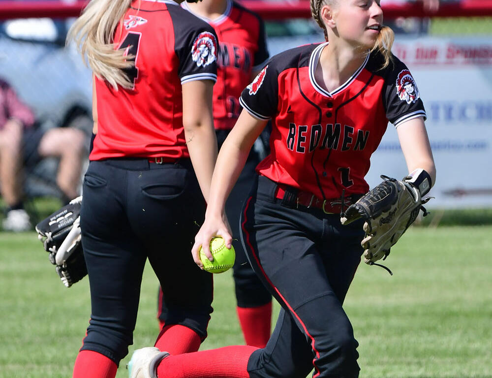 Softball players in red-and-black uniforms on a grassy field; foreground player with glove and a bright green softball ready to throw.