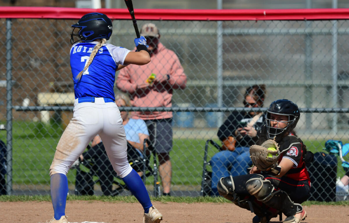 Female softball batter in a blue uniform swings at a pitch while the catcher in black gear crouches behind home plate and a chain-link fence looms in the background.