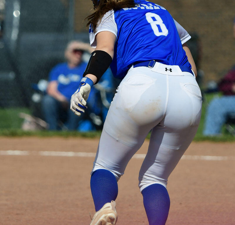 Back view of a softball player in a blue jersey (#8) running on a dirt field, white pants, blue socks, pink cleats, kicking up dust.