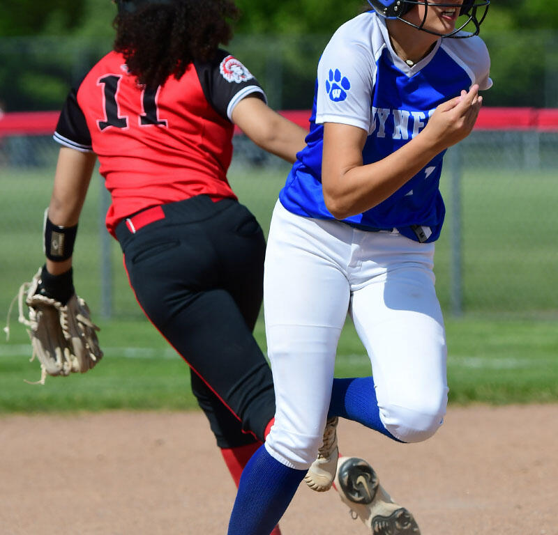 Two softball players sprint on a dirt infield; blue and white uniform leads, red and black uniform pursues behind.